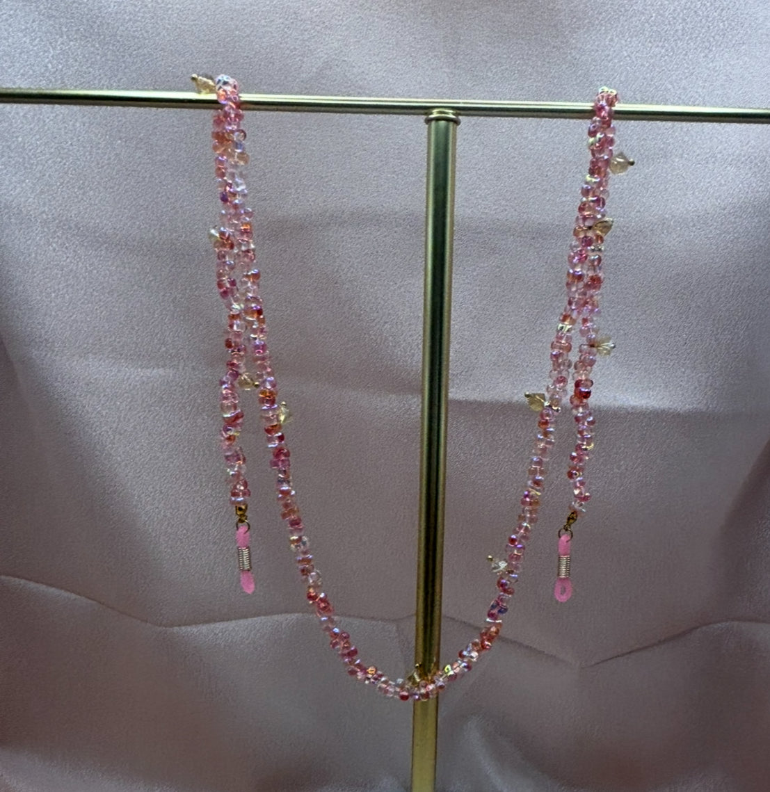 Beaded pink eyeglass chain with iridescent cherry blossom pink seed beads and small gold charms hanging on a gold display stand