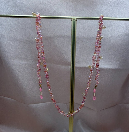 Beaded pink eyeglass chain with iridescent cherry blossom pink seed beads and small gold charms hanging on a gold display stand