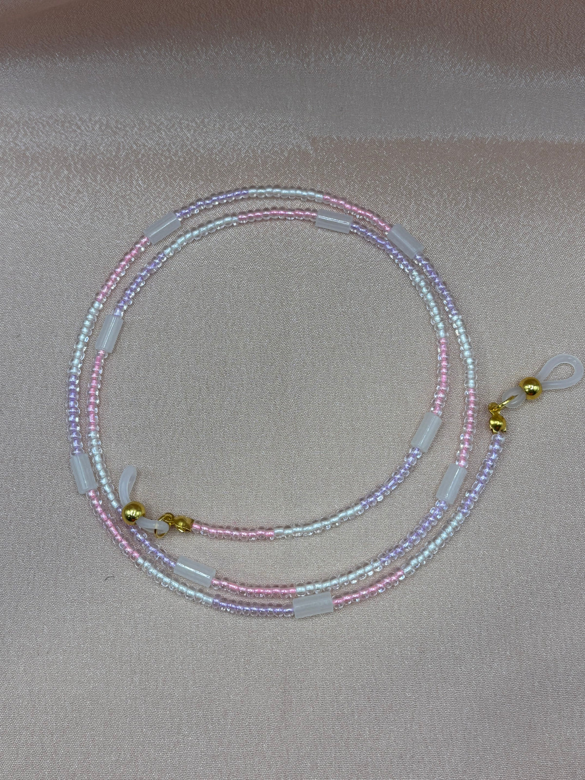A long, coiled beaded eyeglass chain laid flat against a light gray background, showing a mix of clear, white, and subtle pastel beads with metallic clasps at the ends.