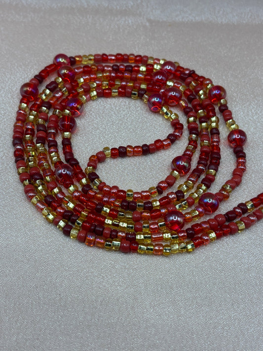 Ruby Flame red and gold beaded eyeglass chain with iridescent crimson accent beads coiled on a pink background.