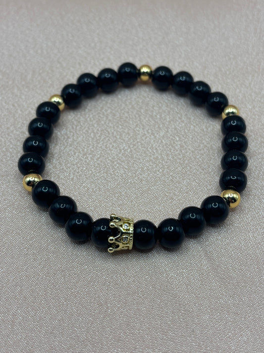 Sovereign handcrafted stretch bracelet featuring sleek polished black stone beads, gold metallic spacer accents, and a detailed crown centerpiece charm by Beadiful Adornments.