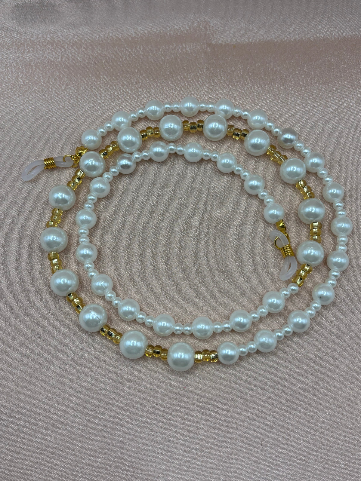Elegant white pearl and gold beaded eyeglass chain coiled on a blush background.